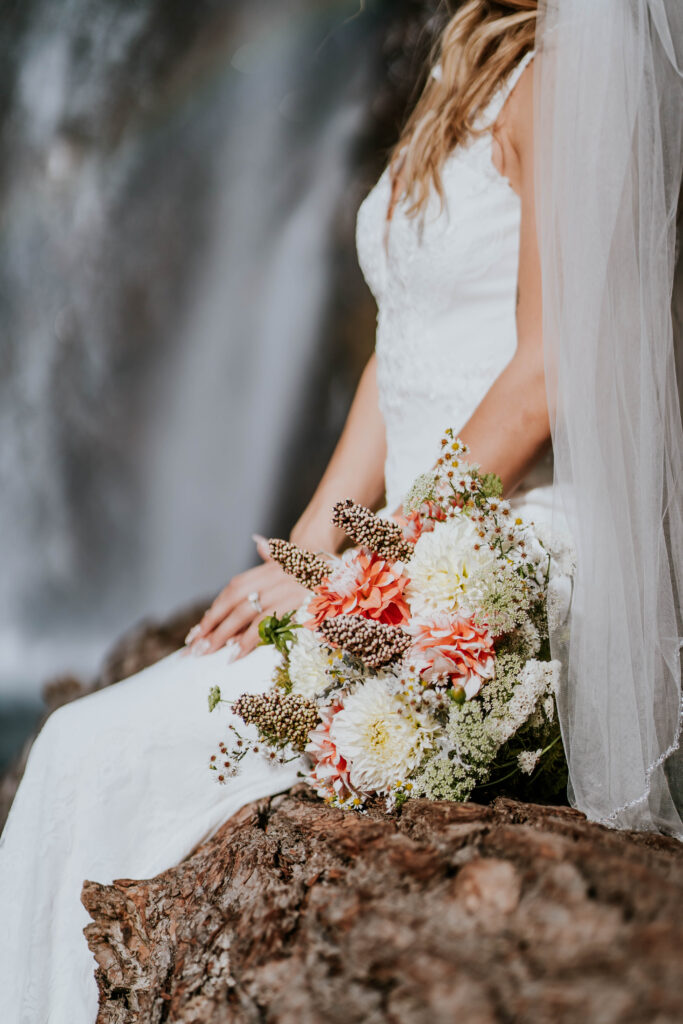 Bride sitting on a log with her bouquet with a waterfall and rainbow in the background