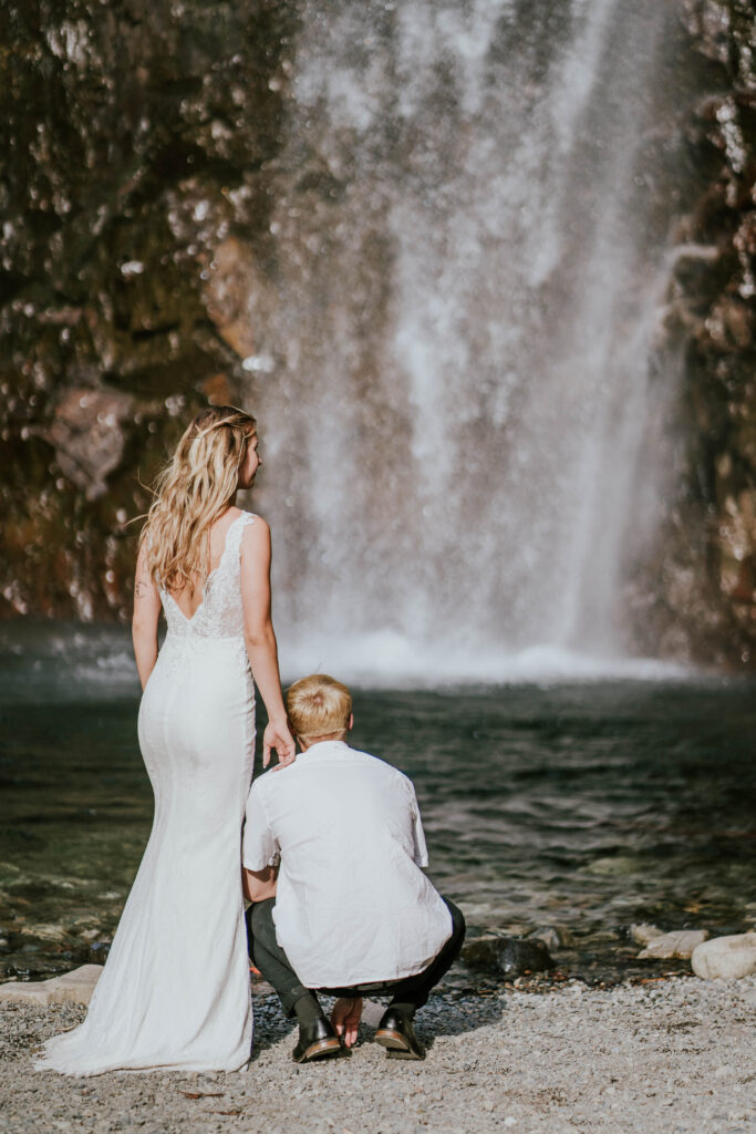 Bride standing and groom squatting facing the waterfall