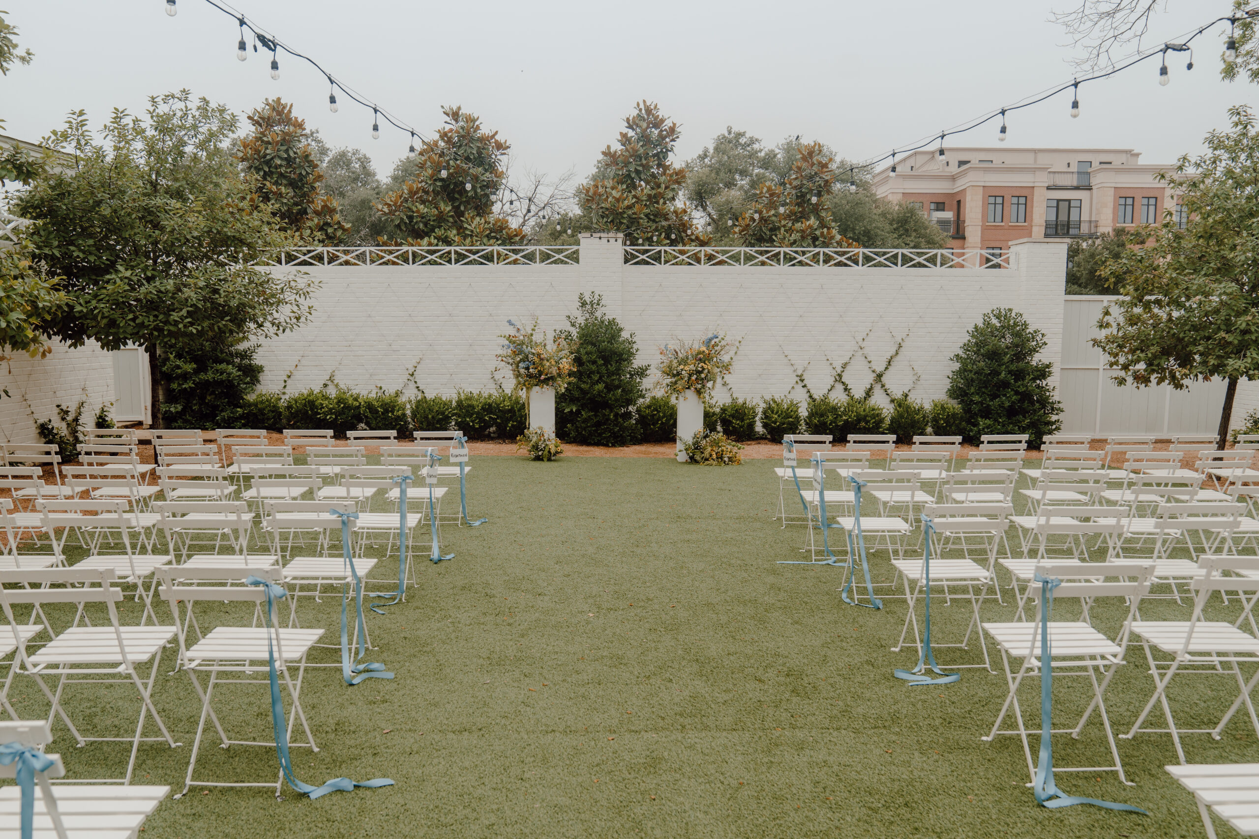 White ceremony chairs set up in rows with string lights overhead and two pillars with florals